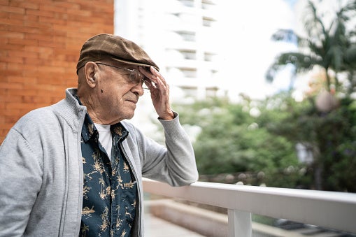 Elderly man standing on his balcony looking longingly off to the side with a very worried express and his hand on his forehead.