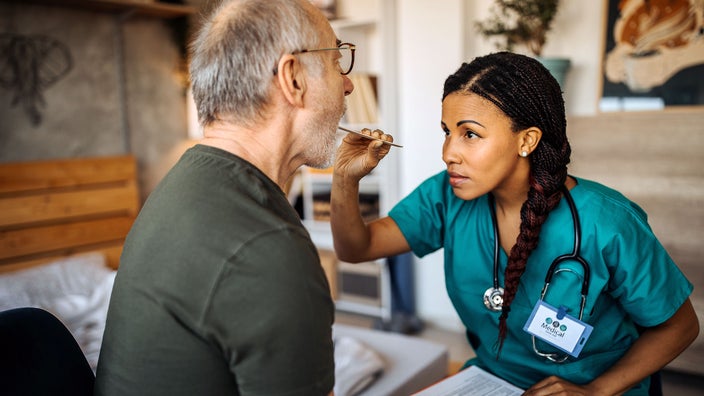 Young nurse in green scrubs checking older male patient's throat with a tongue depressor.