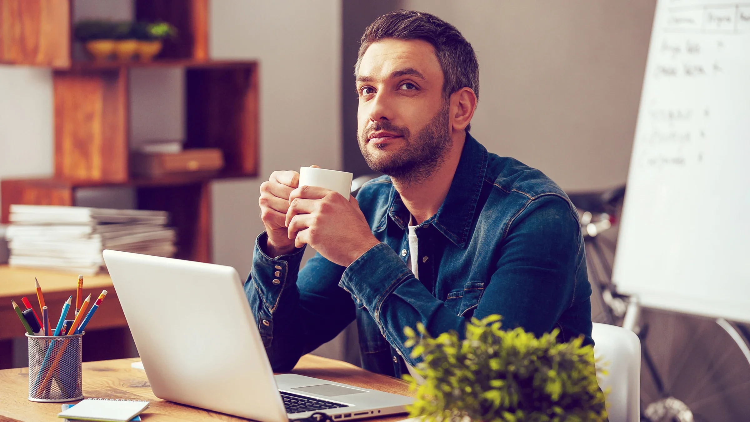 A man daydreams at his desk in his home. He is holding a mug in his hands and is not getting any work done.
g-stockstudio/iStock via Getty Images
