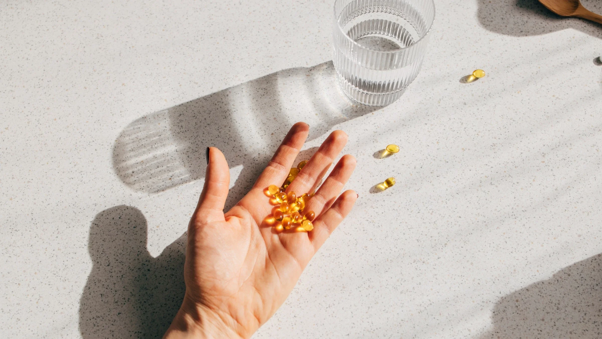 Someone's palm laying flat on a white speckled countertop with a palm full of vitamin D pills with a glass of water next to the hand casting a shadow from the light shining from the right hand side of the frame.