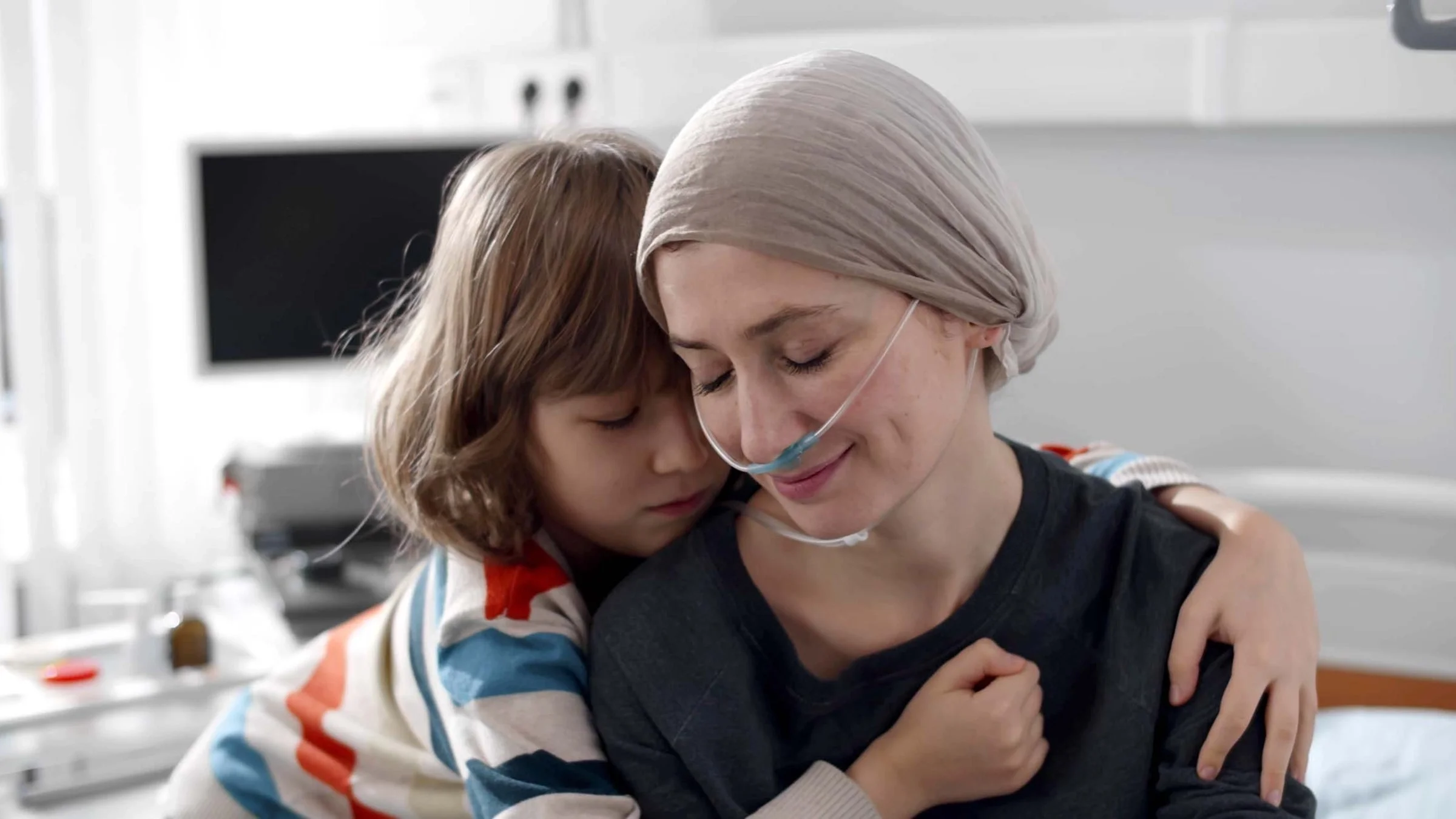 Portrait of a young child hugging their mother in the hospital room. The mother is wearing a tan headscarf and had oxygen tubes in her nose.