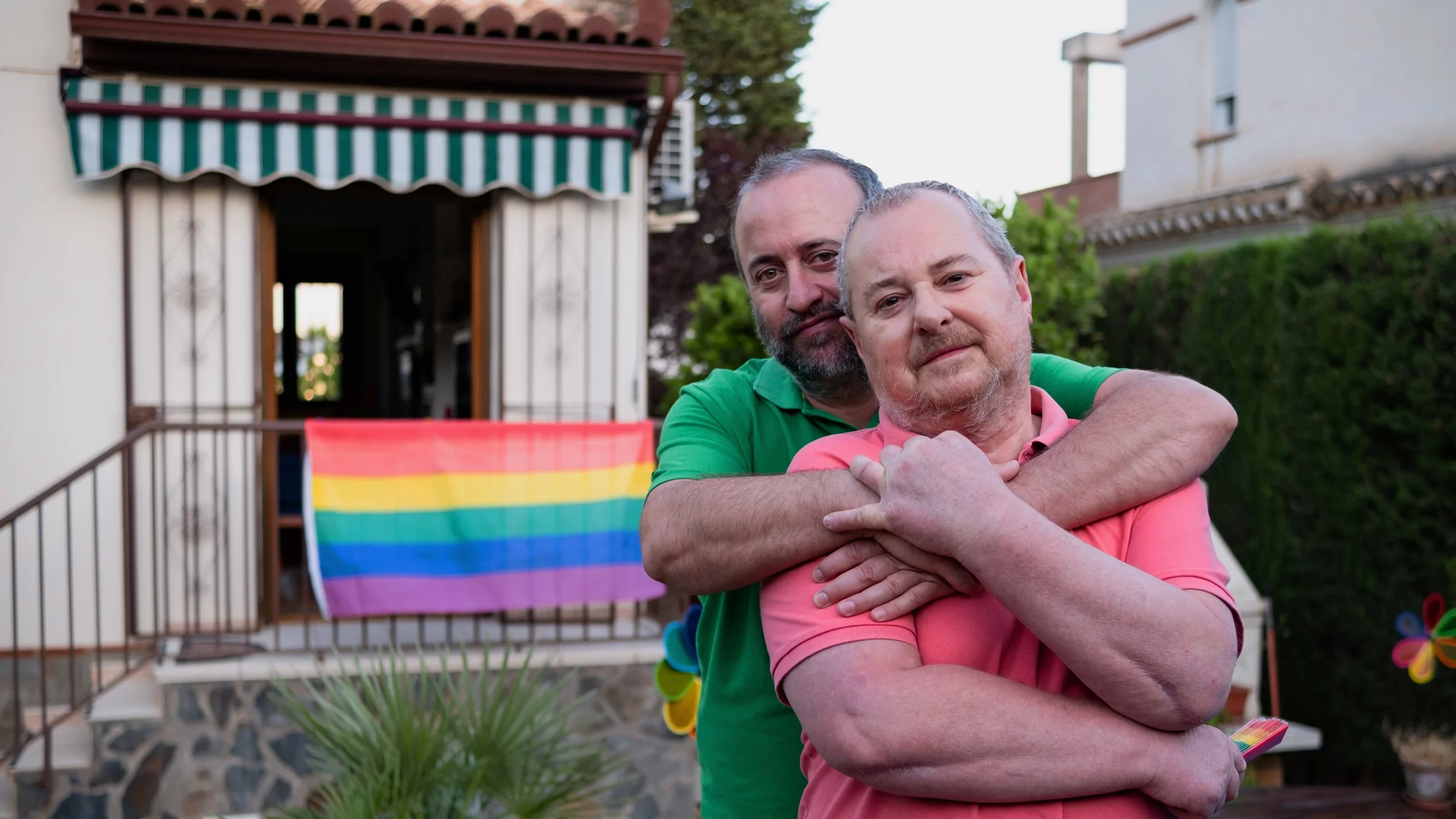 An older gay couple hugging outside their home with a rainbow flag.