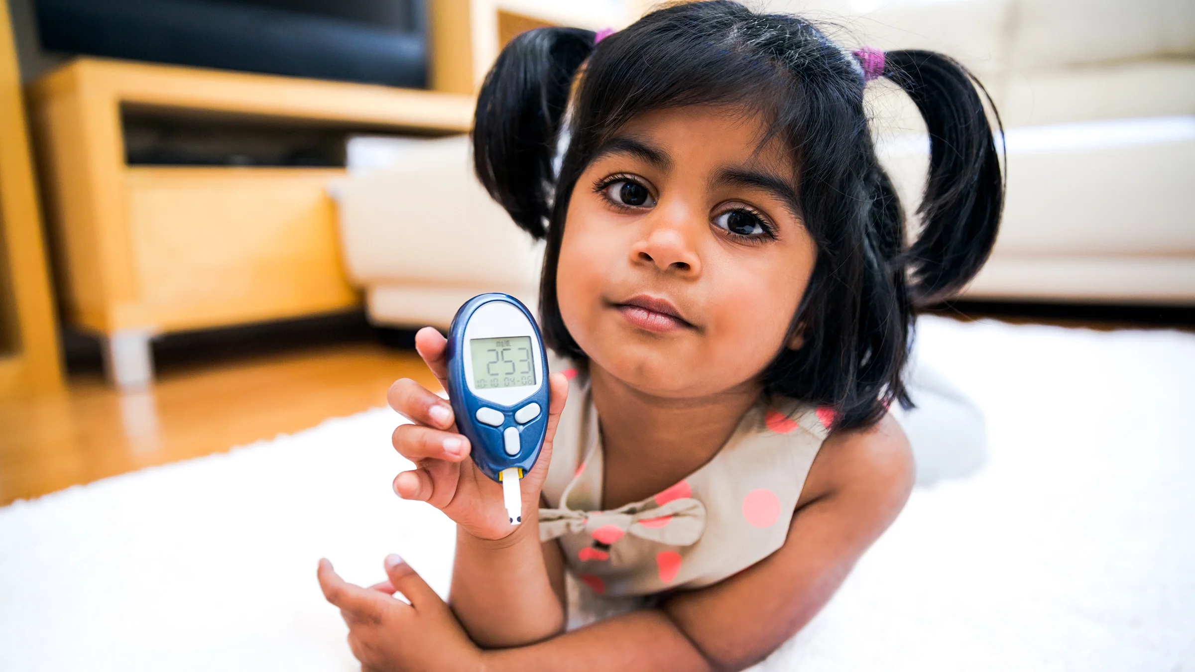 Una niña pequeña y linda con colitas acostada sobre su vientre en el medio de la sala mostrando a la cámara su glucómetro de la sangre.
