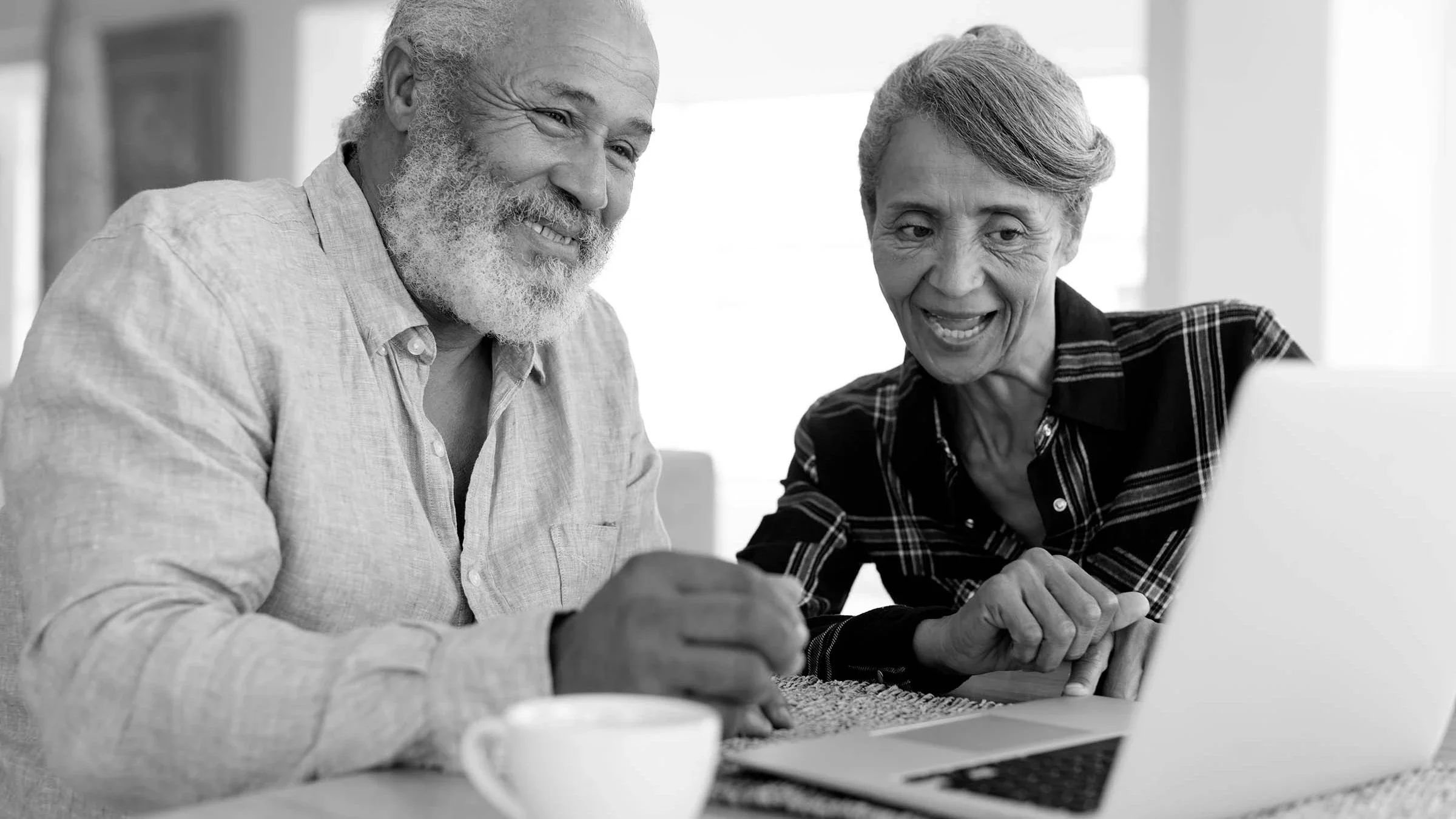Elderly couple looking at something on their laptop at the dining table. There is a coffee cup next to the laptop on the table. Both people are talking and smiling.