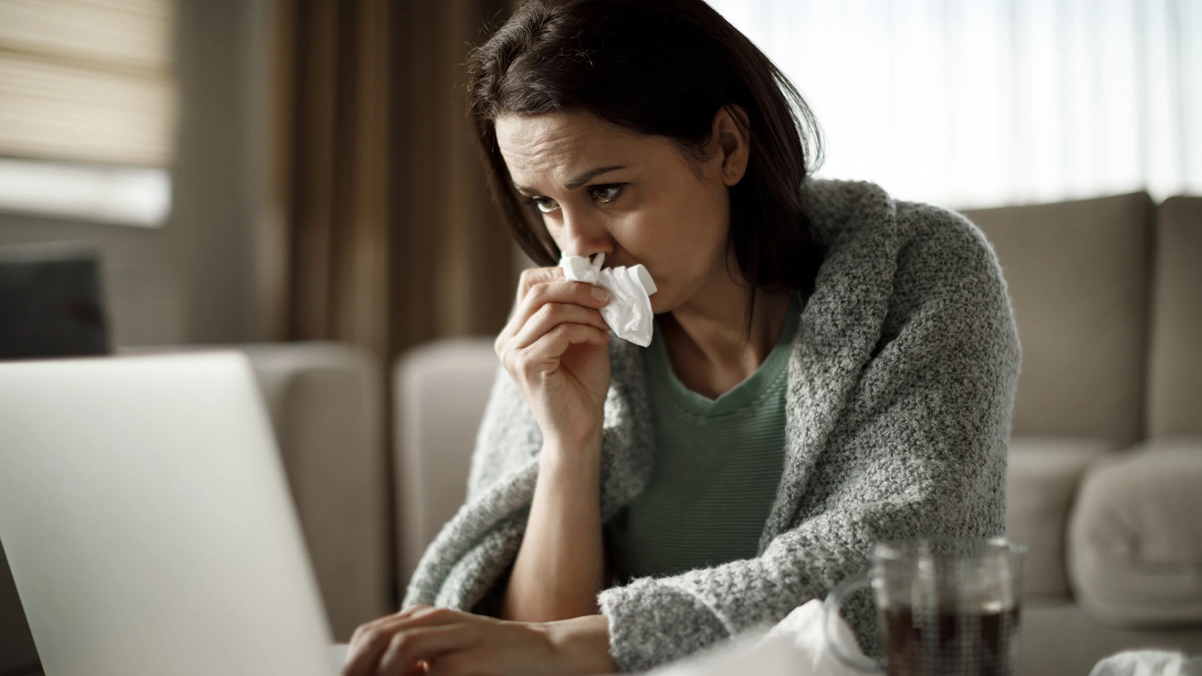 A woman covers her mouth with a tissue as she uses her laptop.
