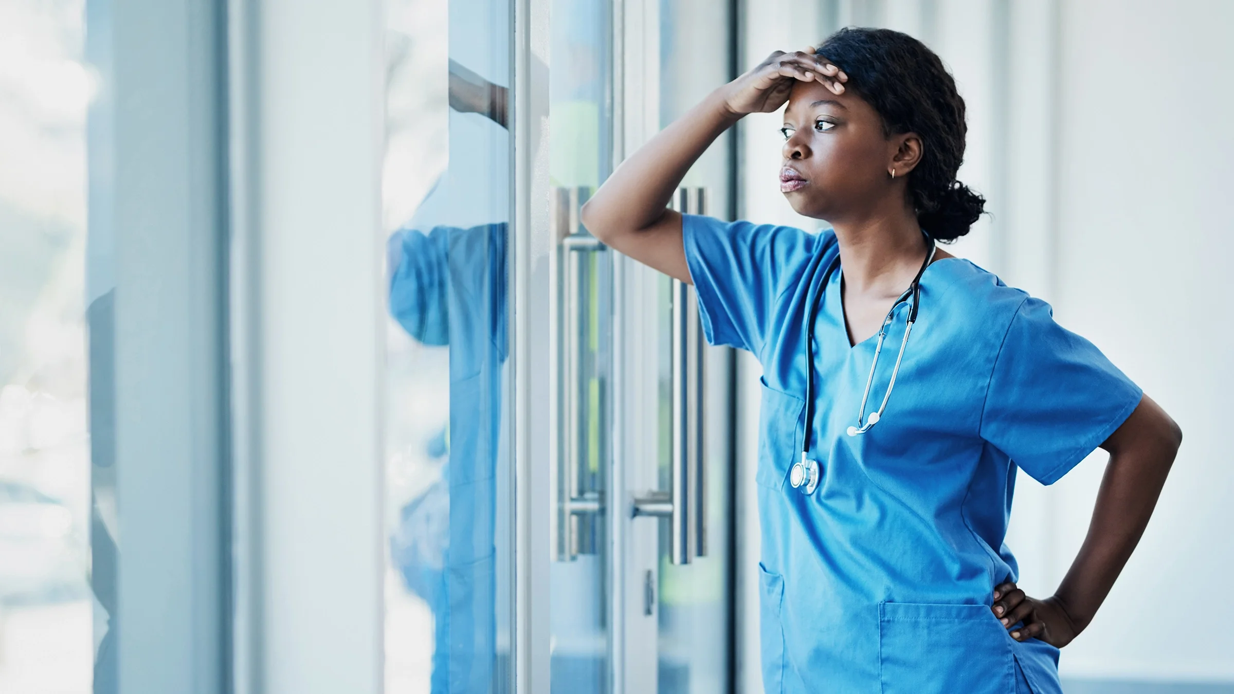 Nurse taking a break by the hospital window. She looks extremely stressed with her hand on her head.