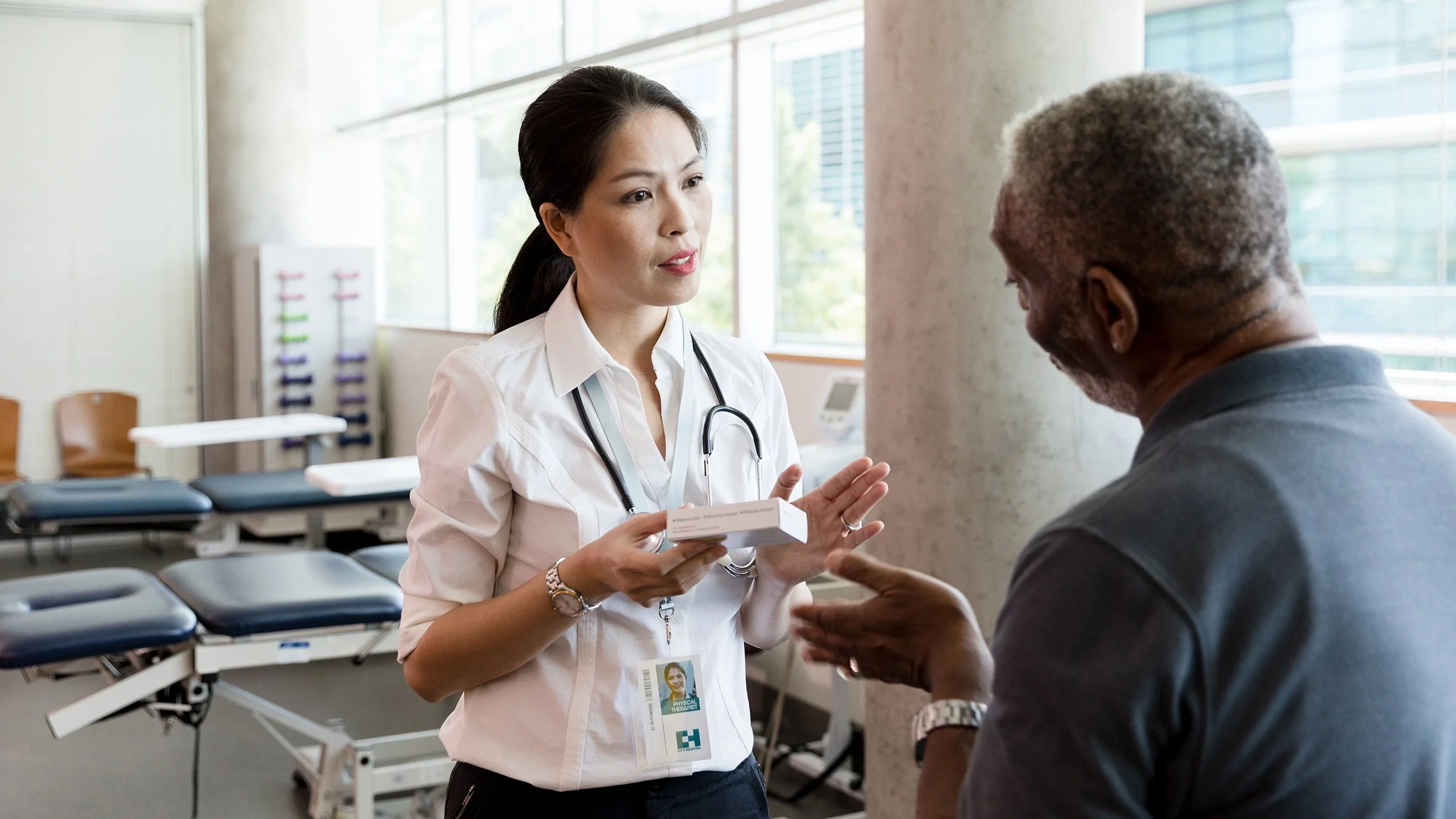 Doctor reviewing a prescription in a white box with a patient. The patient is sitting on an exam table with their back to the camera.