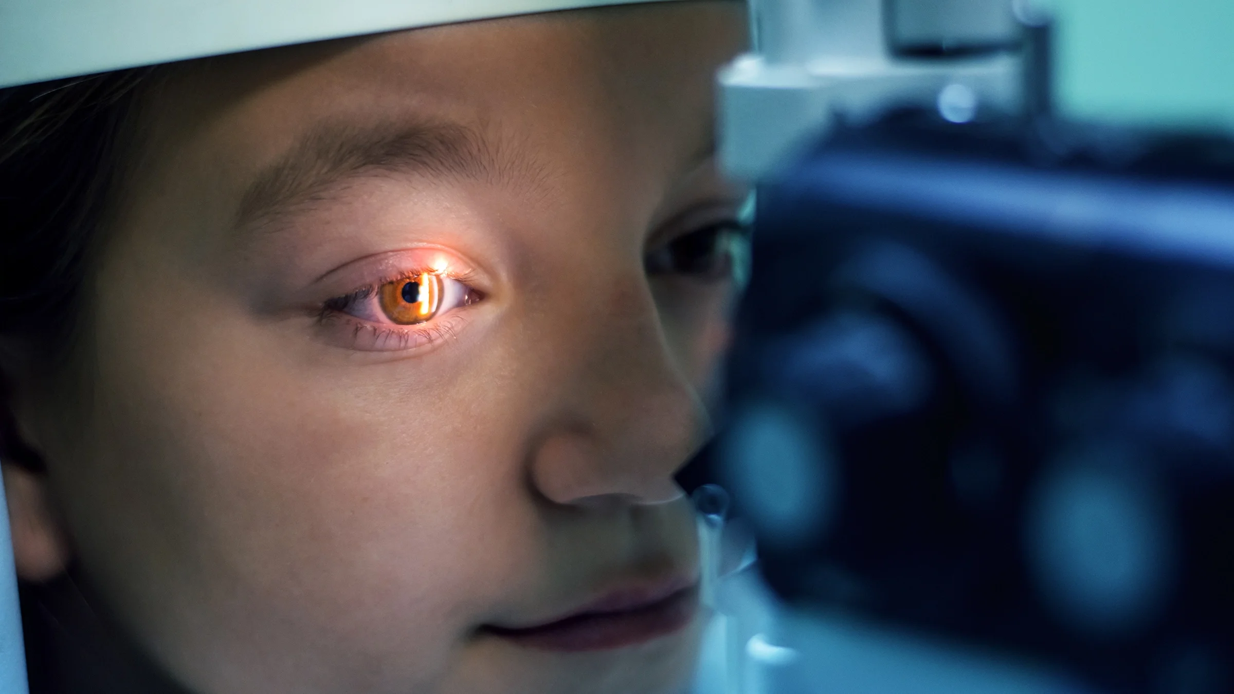 Close-up of a woman having an eye exam. There is a bright white light shining into her eye as she rests her chin on the exam equipment.