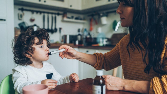 Mother giving her toddler son a syringe ful of cough syrup at the kitchen table.