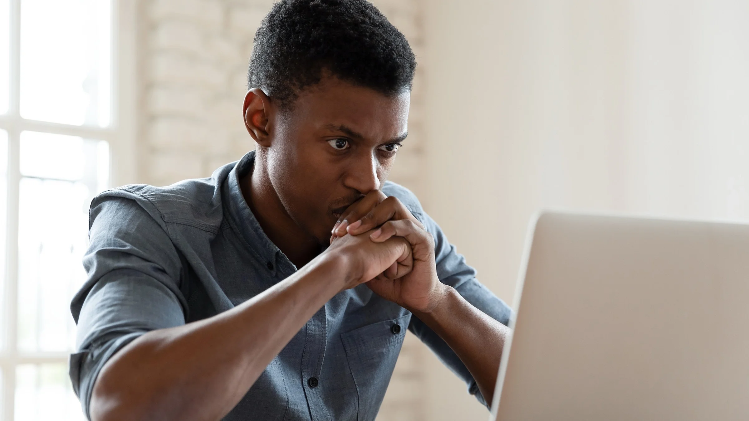 A young man is researching something on the internet on his laptop. He looks extremely concerned with his face resting on his fists.