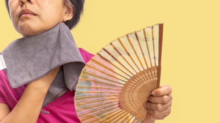 Cropped shot of a woman experiencing a hot flash. She has a towel and a fan in hand to cool her down. The background is a plain yellow studio background.