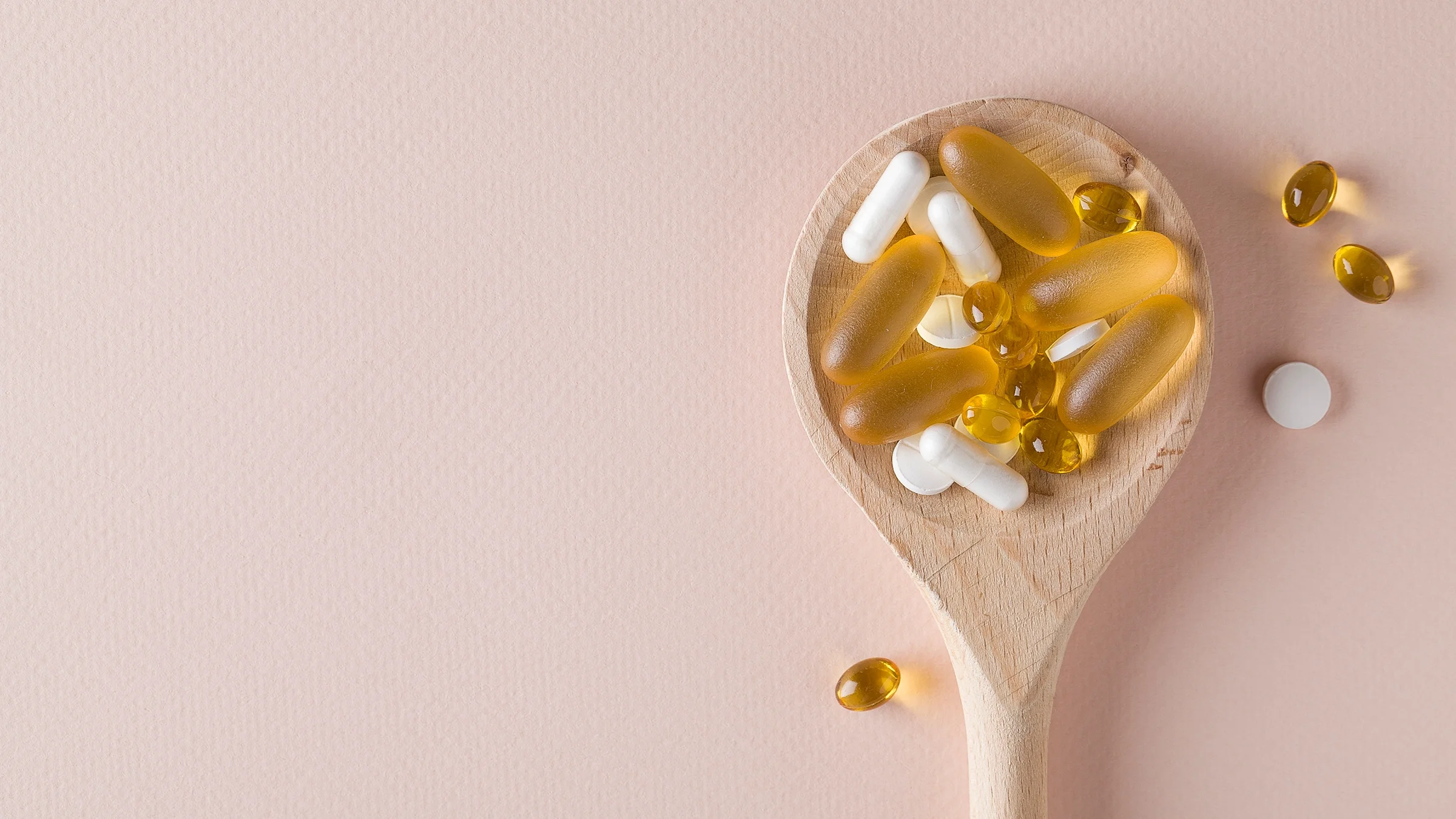 Wooden spoon filled with various supplements on a light pink background.