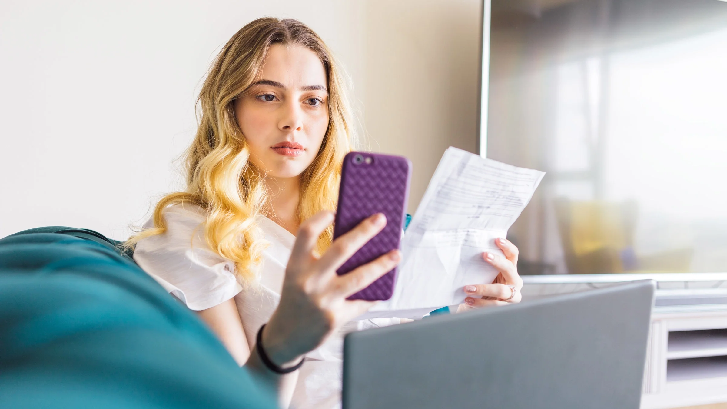 A person with a surprised expression, looking at a paper bill and their phone.