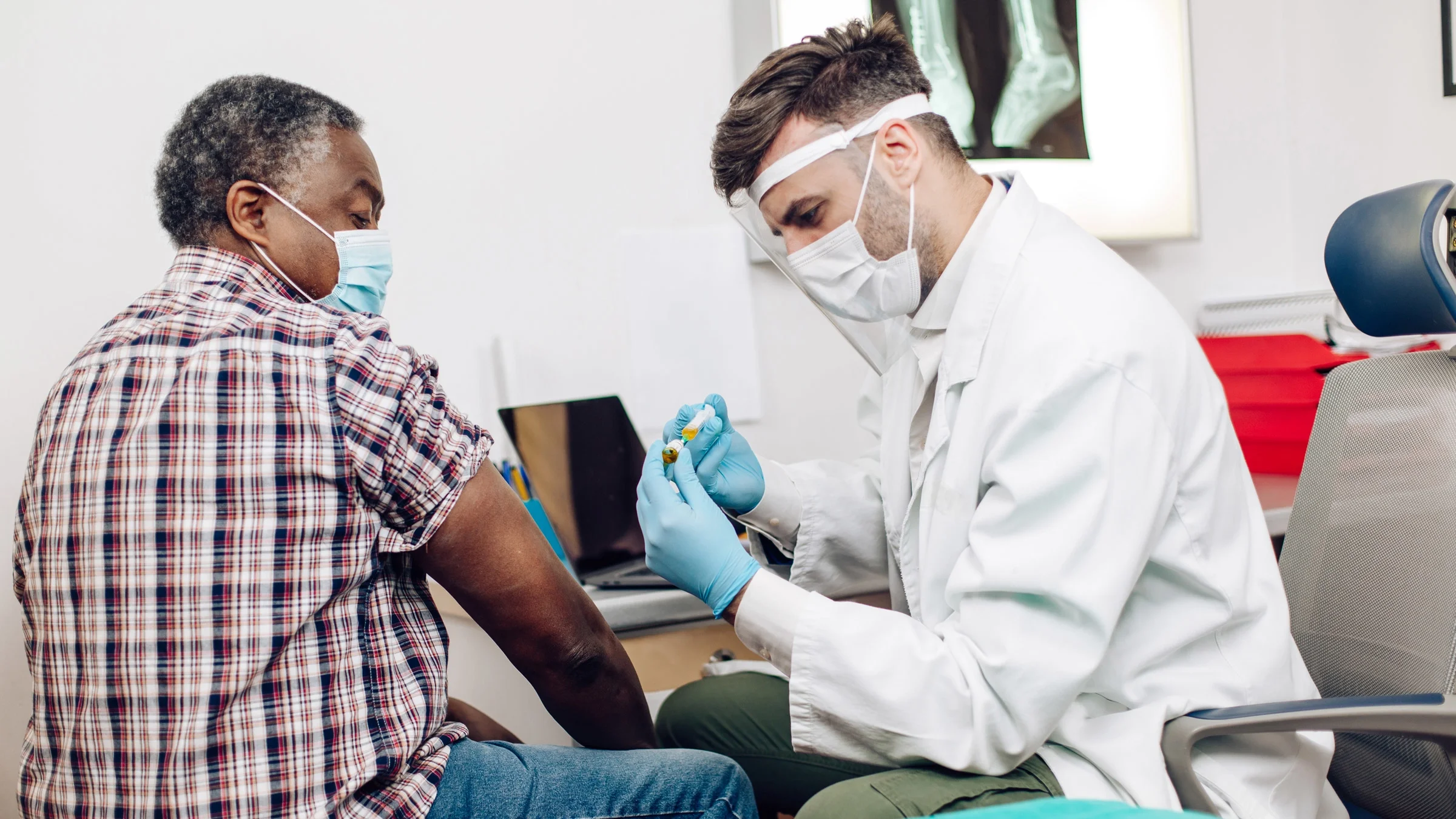 Older man with his sleeve rolled up, sitting next to a male provider who is preparing an injection to give to the patient. 