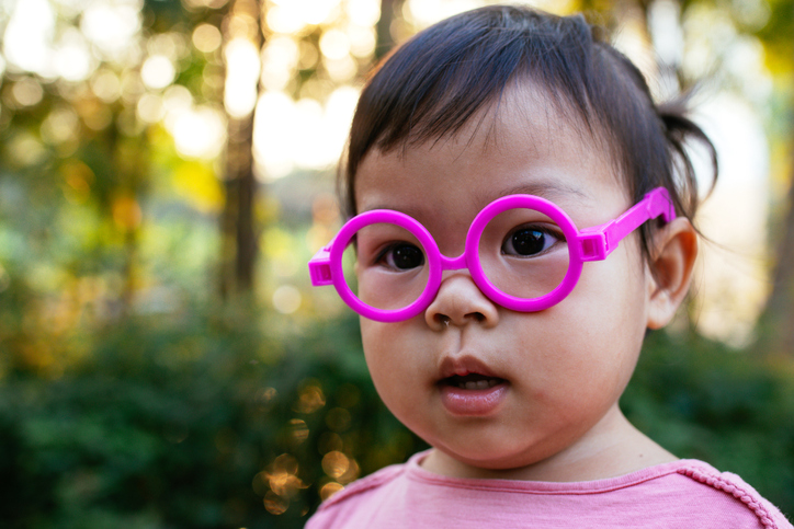 Portrait of the cutest baby girl with her mouth agape wearing pink plastic glasses outside among the trees.