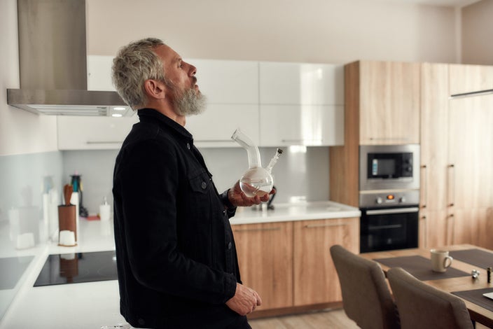 Middle aged man with gray hair and beard smoking out of bong in his kitchen.