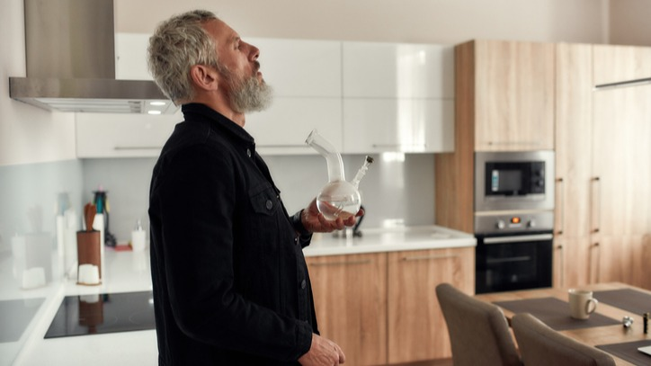 Middle aged man with gray hair and beard smoking out of bong in his kitchen.