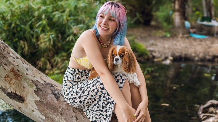 A person with colorful hair cuddling with their dog by a lake.
