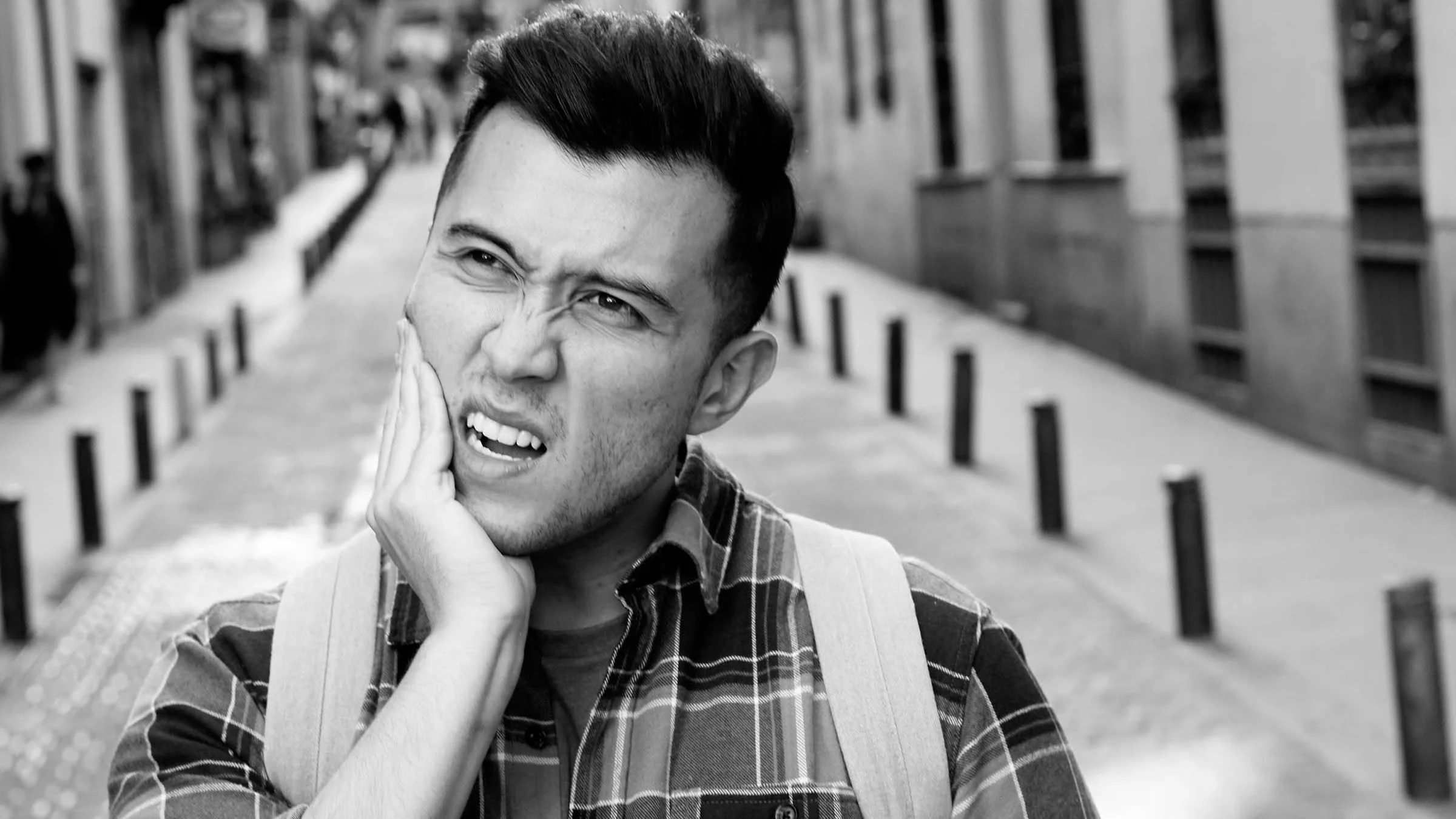 Black and white portrait of a man grabbing his jaw in pain. He is walking down the street with a backpack on.