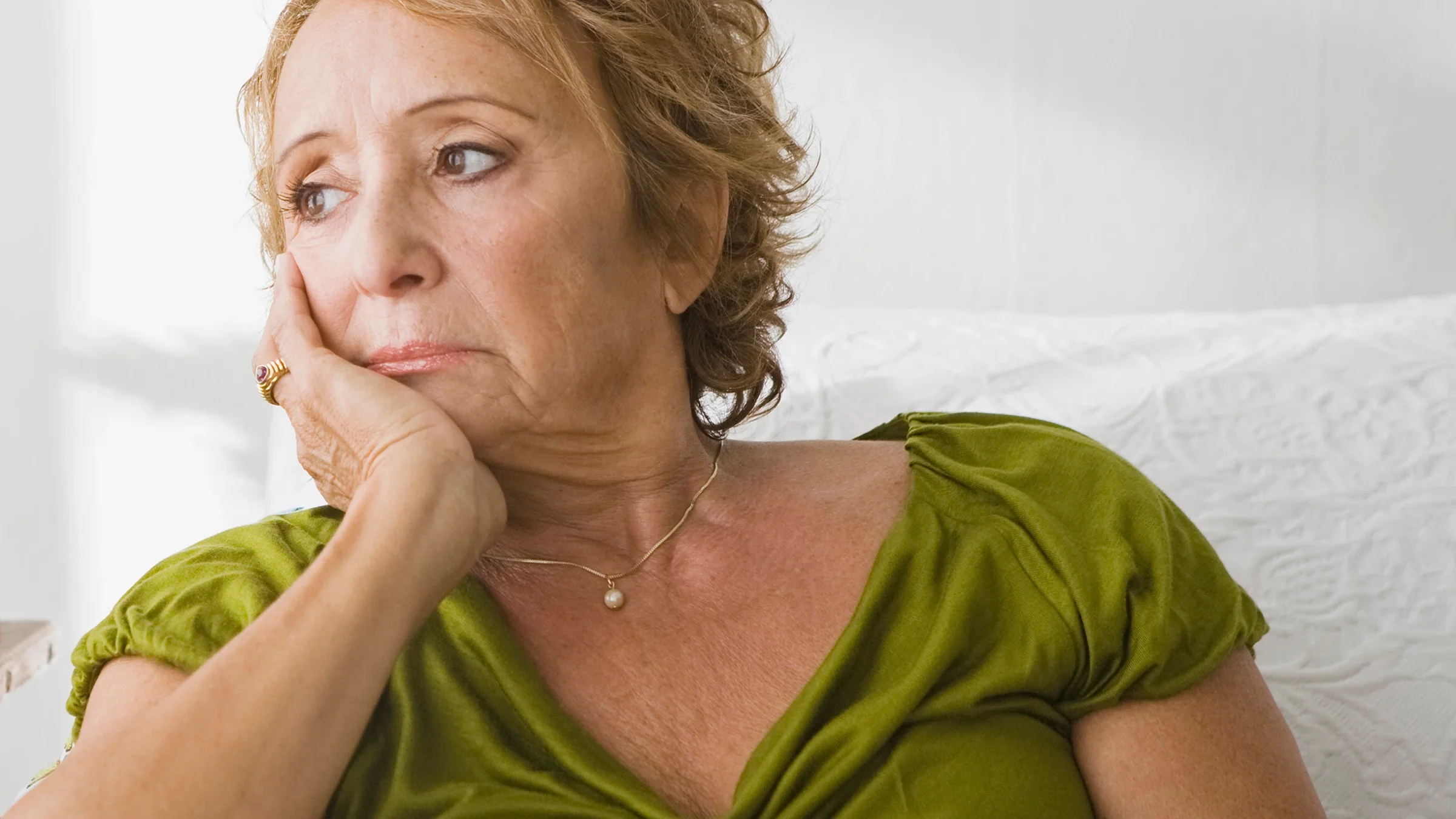Close-up on an older woman looking sad and resting her cheek on her hand. She is wearing a green blouse.