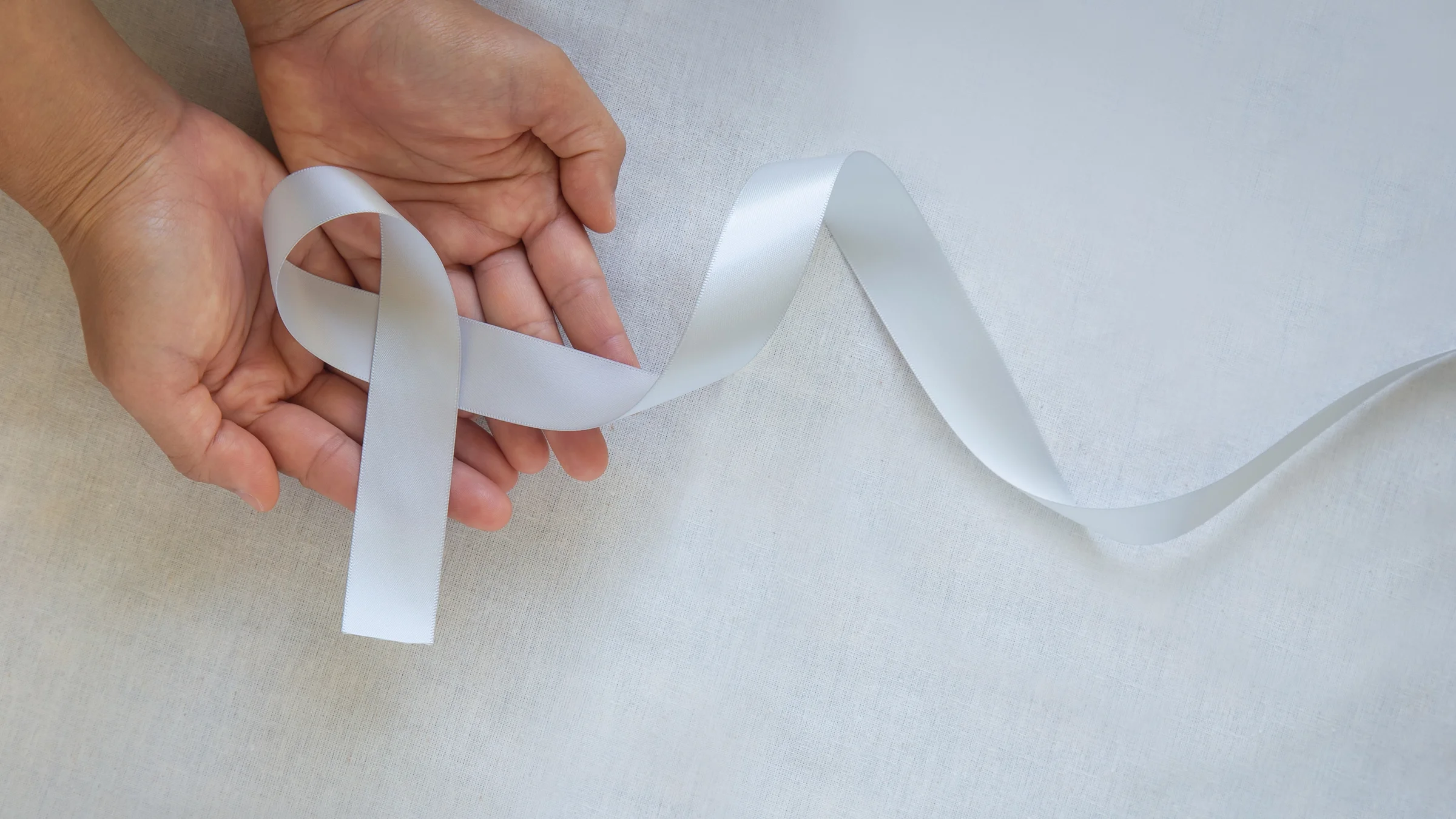 A hand is holding a silver cause ribbon on a light background.