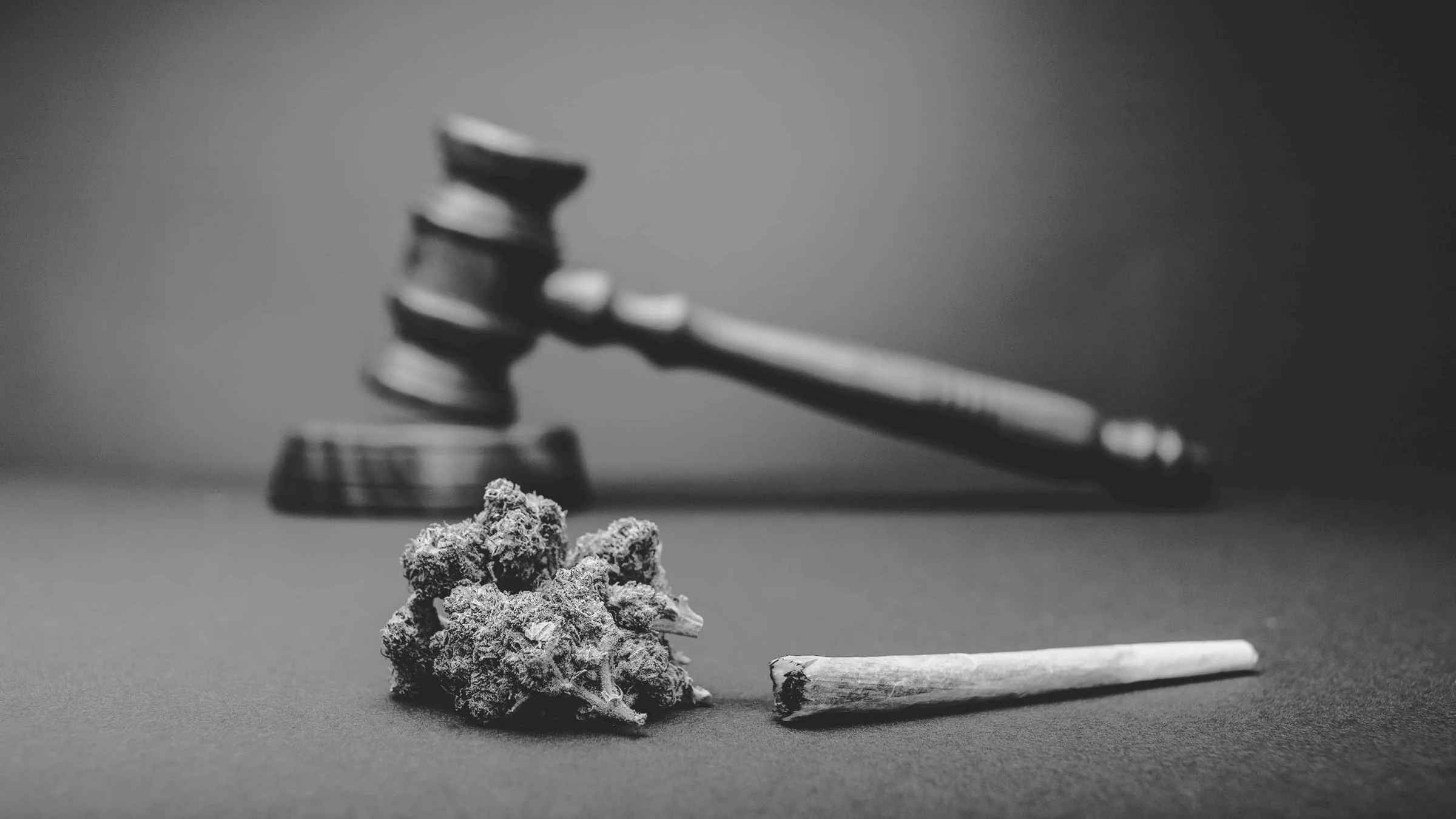 Black and white photo of marijuana bud and a joint on a gray background with a wooden judicial hammer in the background.