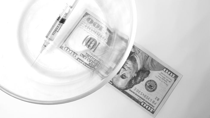 Black and white image of a vaccine syringe in a clear bowl covering a hundred dollar bill on a white background.