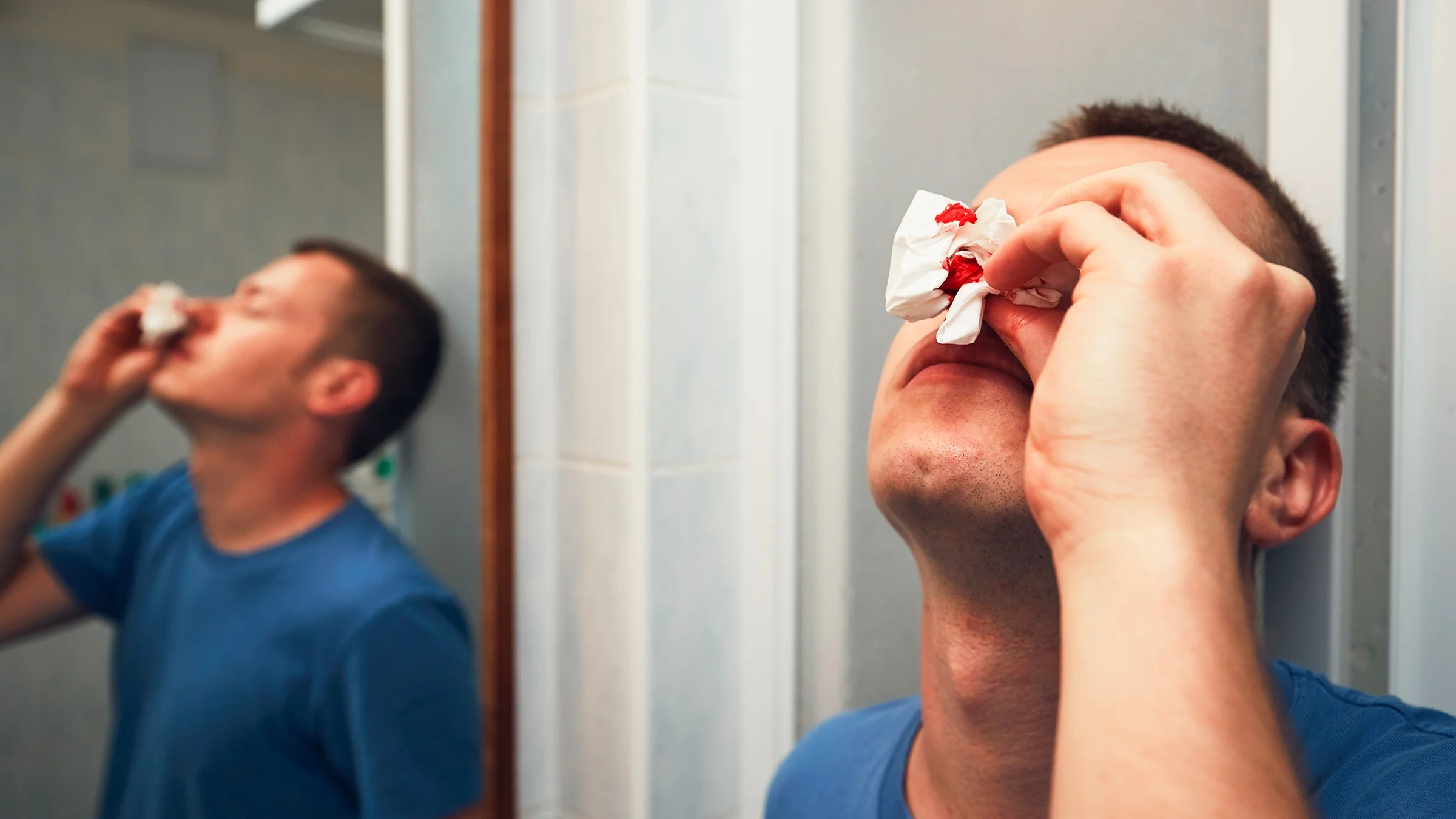 An adult with a nose bleed is tilting their head back in the bathroom.