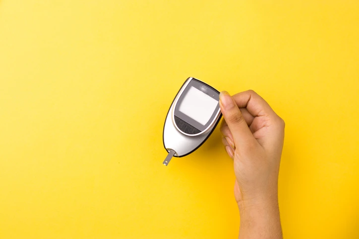 Hand holding a glucometer on a yellow background.
