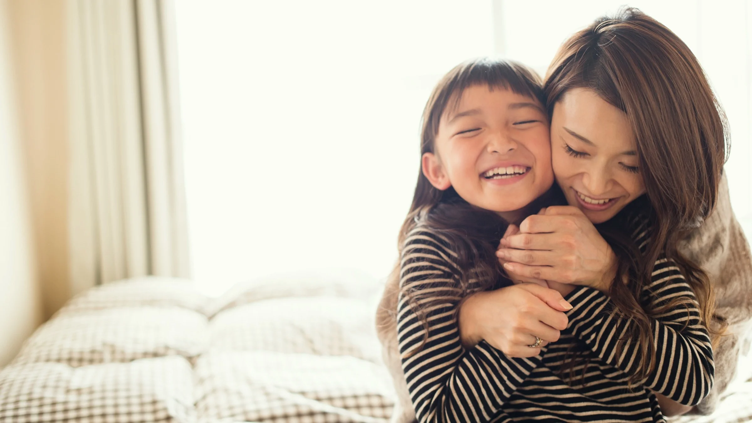 A mother is hugging her daughter from behind. Both are smiling and laughing.