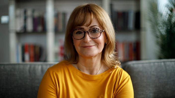 Portrait of an older woman wearing a bright yellow long-sleeve shirt and sitting on a couch.