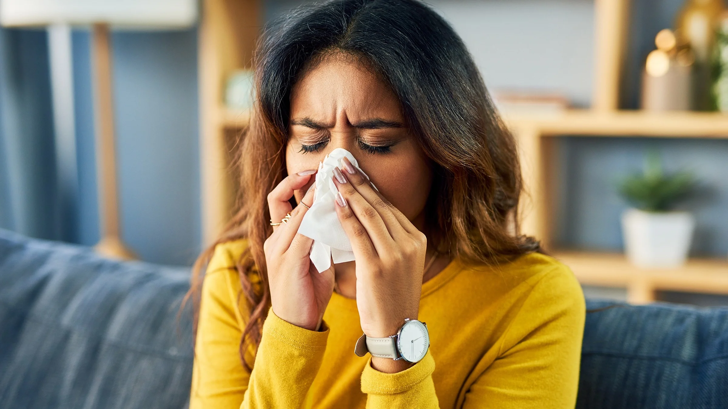 Portrait of a woman blowing her nose while sitting on her couch. She is wearing a yellow long sleeve shirt.