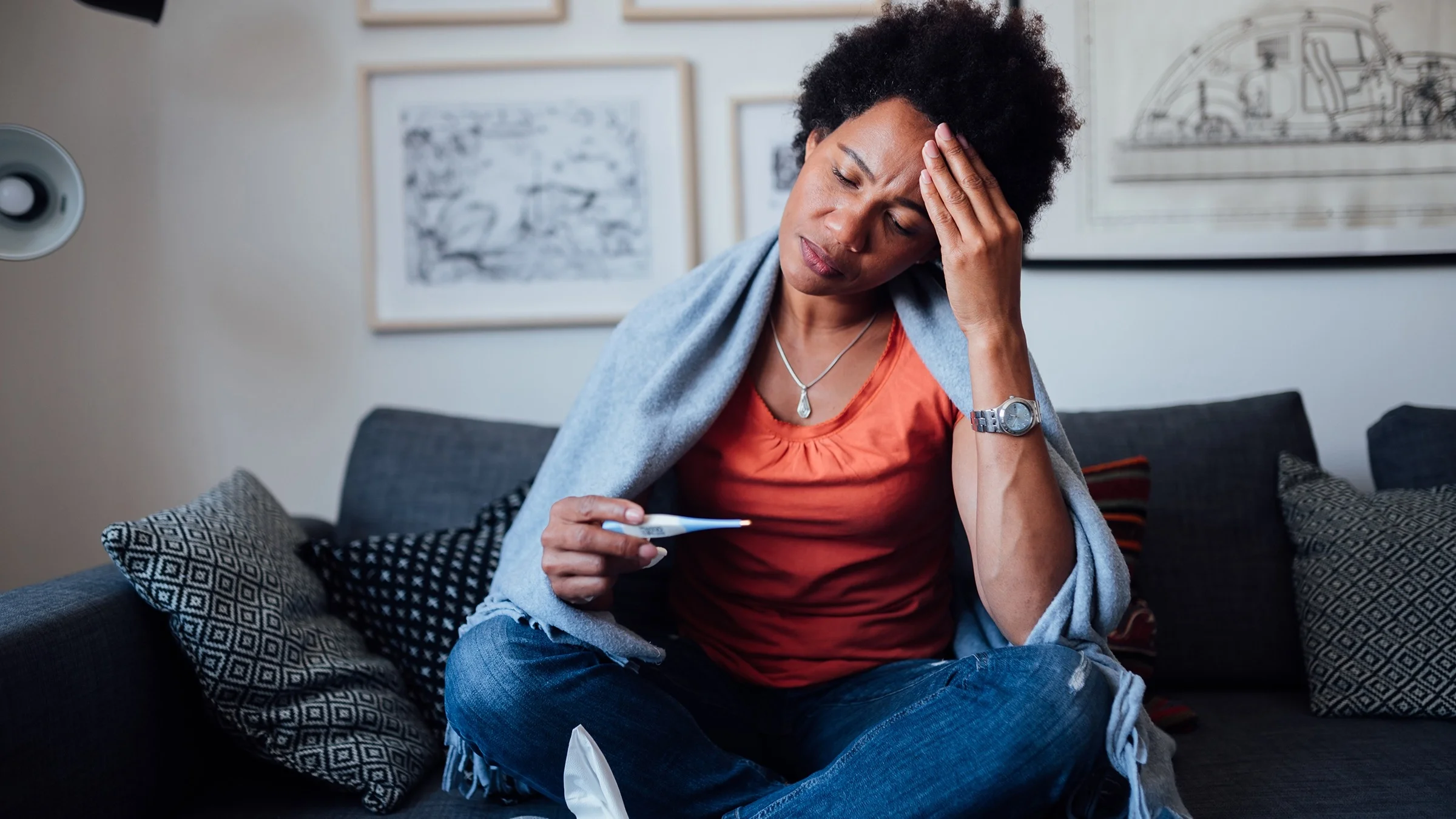 Young woman sitting cross legged on her couch taking her temperature with a thermometer. She has her hand on her head with a concerned look on her face.