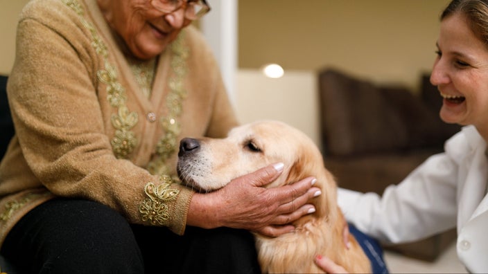 An older woman and her caregiver are with a service dog at home.