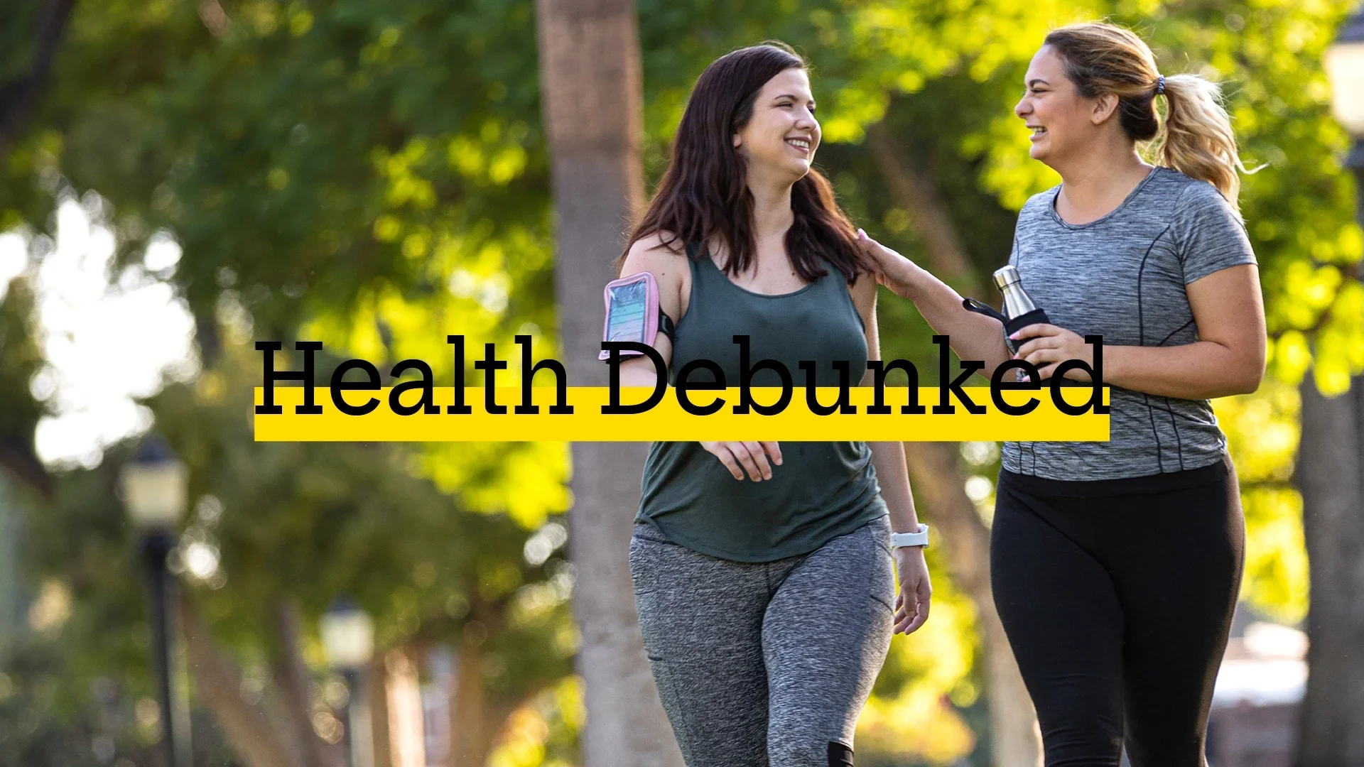 Two friends going for a walk through the park. They are both wearing work out clothes. There is added text on top reading “Health Debunked” with a yellow highlight bar behind it.