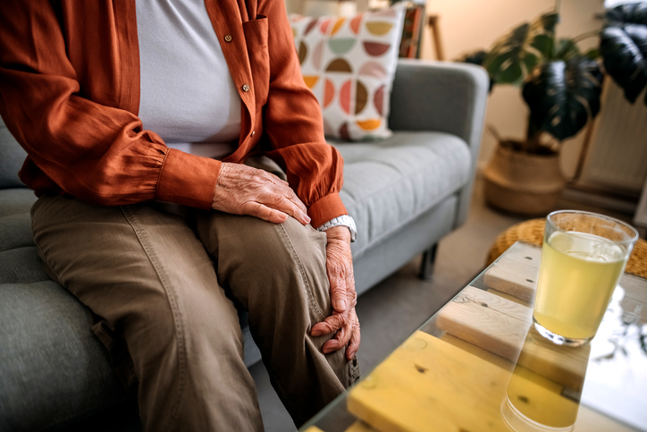 Close-up cropped shot of an elderly woman sitting on the couch reaching for her sore knee.