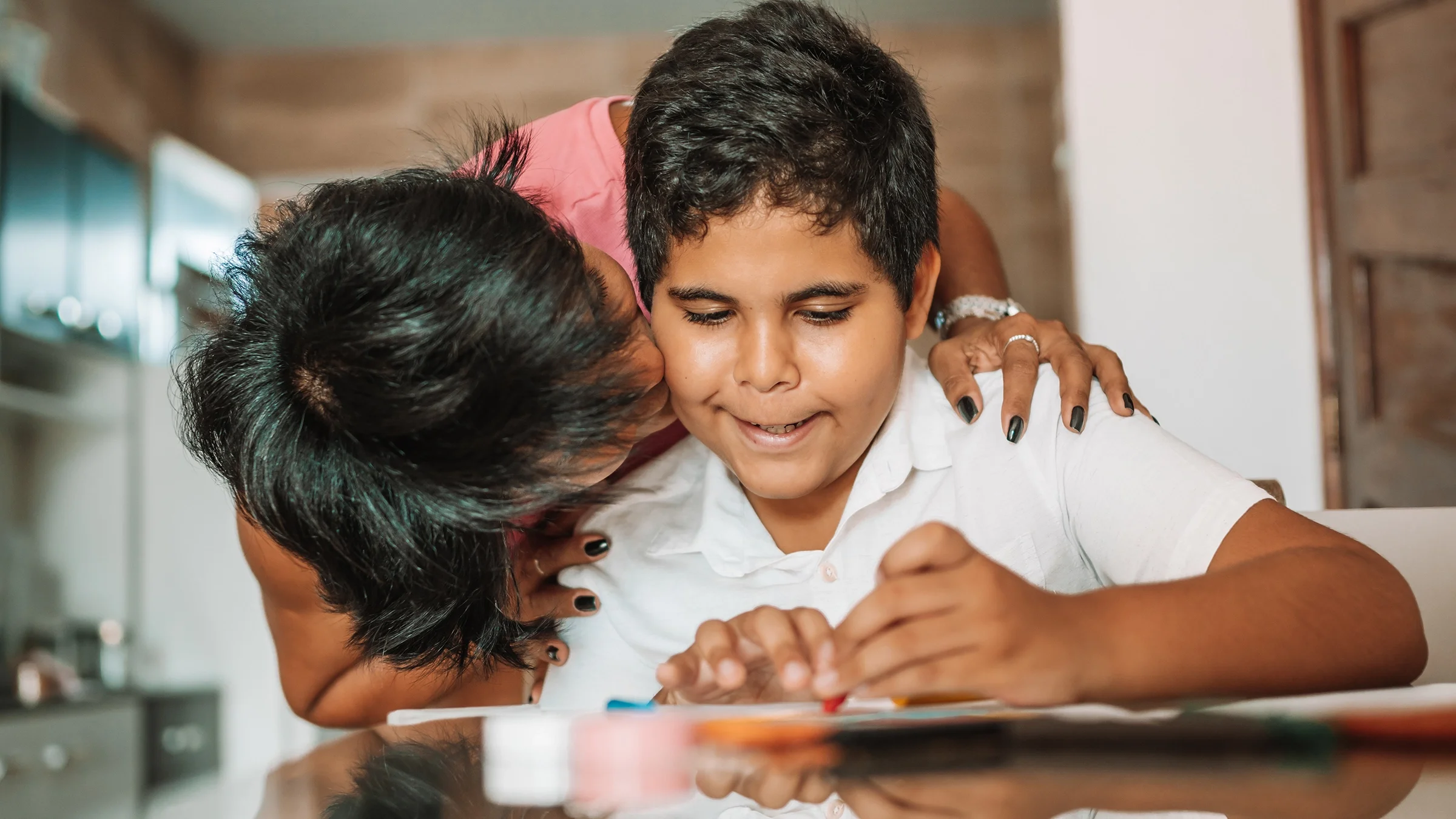 A mother is bending down to kiss her son on the cheek as he does crafts at home.