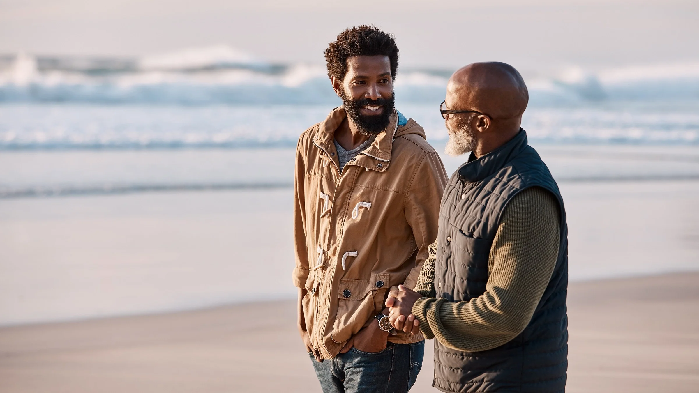 Adult son and father walking along the beach during the winter. They are both wearing sweaters and jackets that are neutral colors.