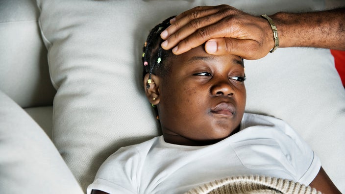Young child sick in bed as a parent uses their hand to test the child’s temperature.