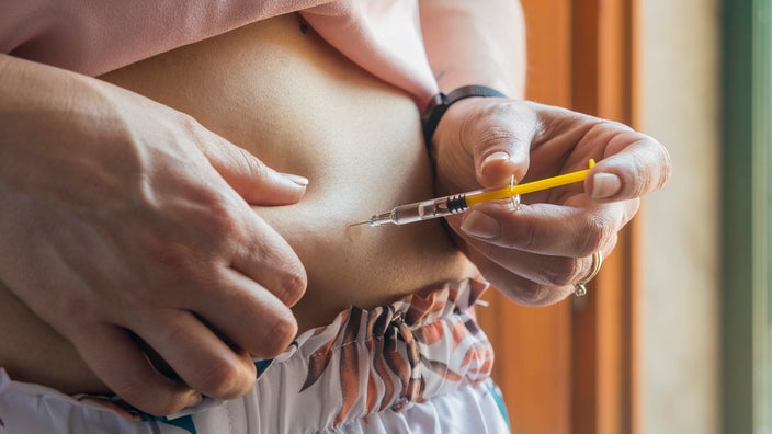 Close-up of a person injecting themselves in the abdomen with a yellow syringe. They have floral pjamas on.