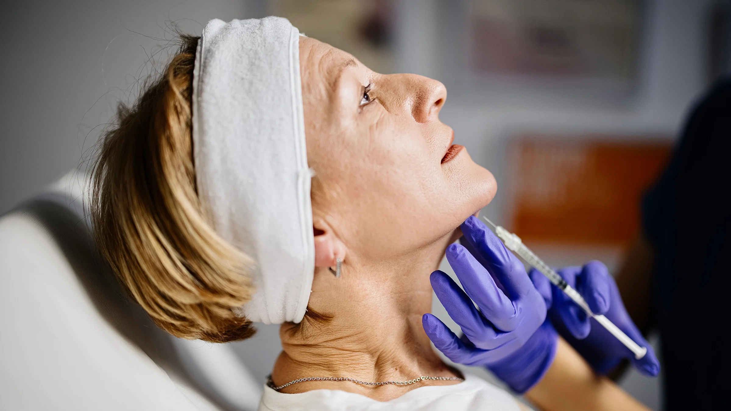 Older woman getting prepped with an injection for a double chin surgery. She has white headband holding her hair back while the nurse administers the shot.