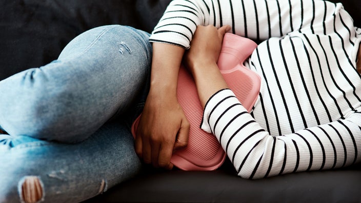 A woman holds a hot water bottle against her stomach on the sofa at home. This is one of several at-home remedies to get rid of period cramps fast. 