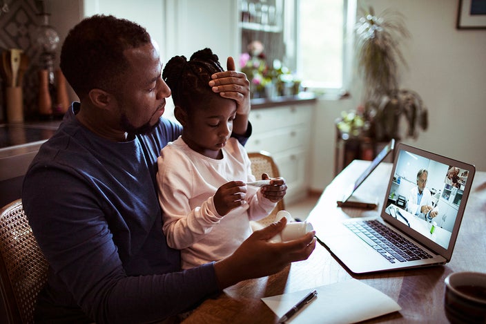 A father holding with his daughter on his lap at the kitchen table talking to the doctor on a telehealth visit. He is feeling her forehead as he reads the thermometer.