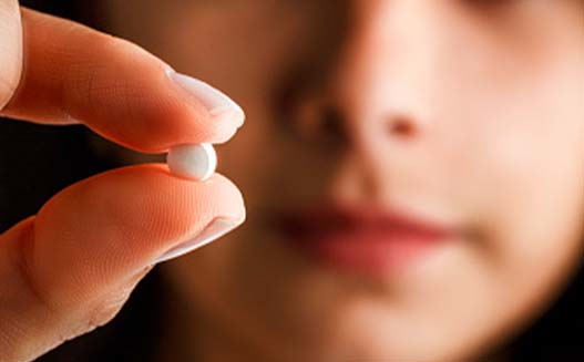Close-up of a young Hispanic woman holding up a single round white pill.