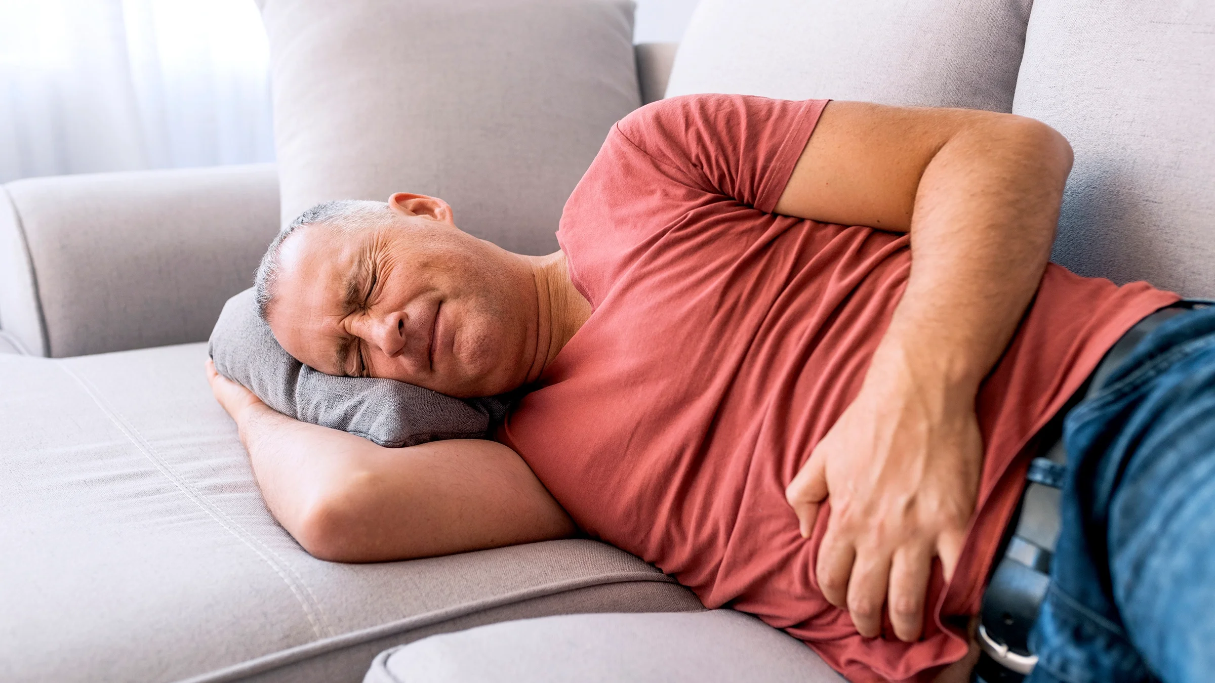 Older man with salt and pepper hair in severe pain laying on the couch clutching his stomach.