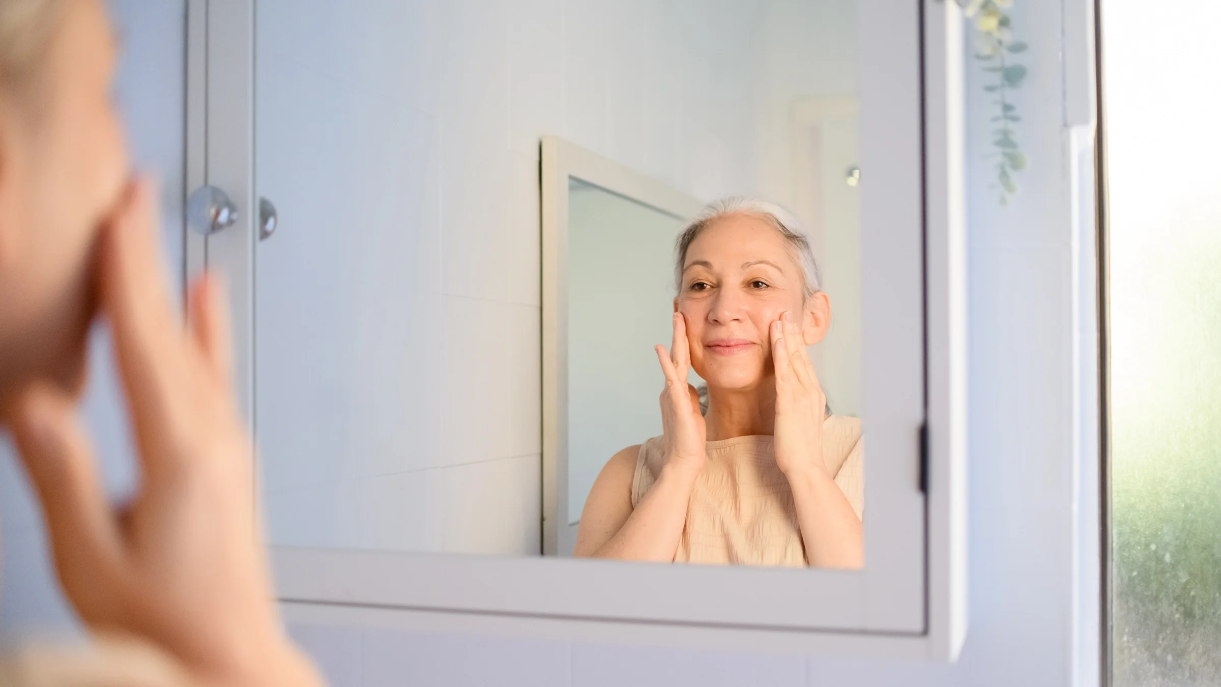 A woman applies cream to her face in front of a mirror.