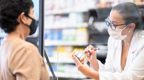 Pharmacist wearing a face mask reviewing a patient's prescription with them over the counter through a plexiglass barrier.