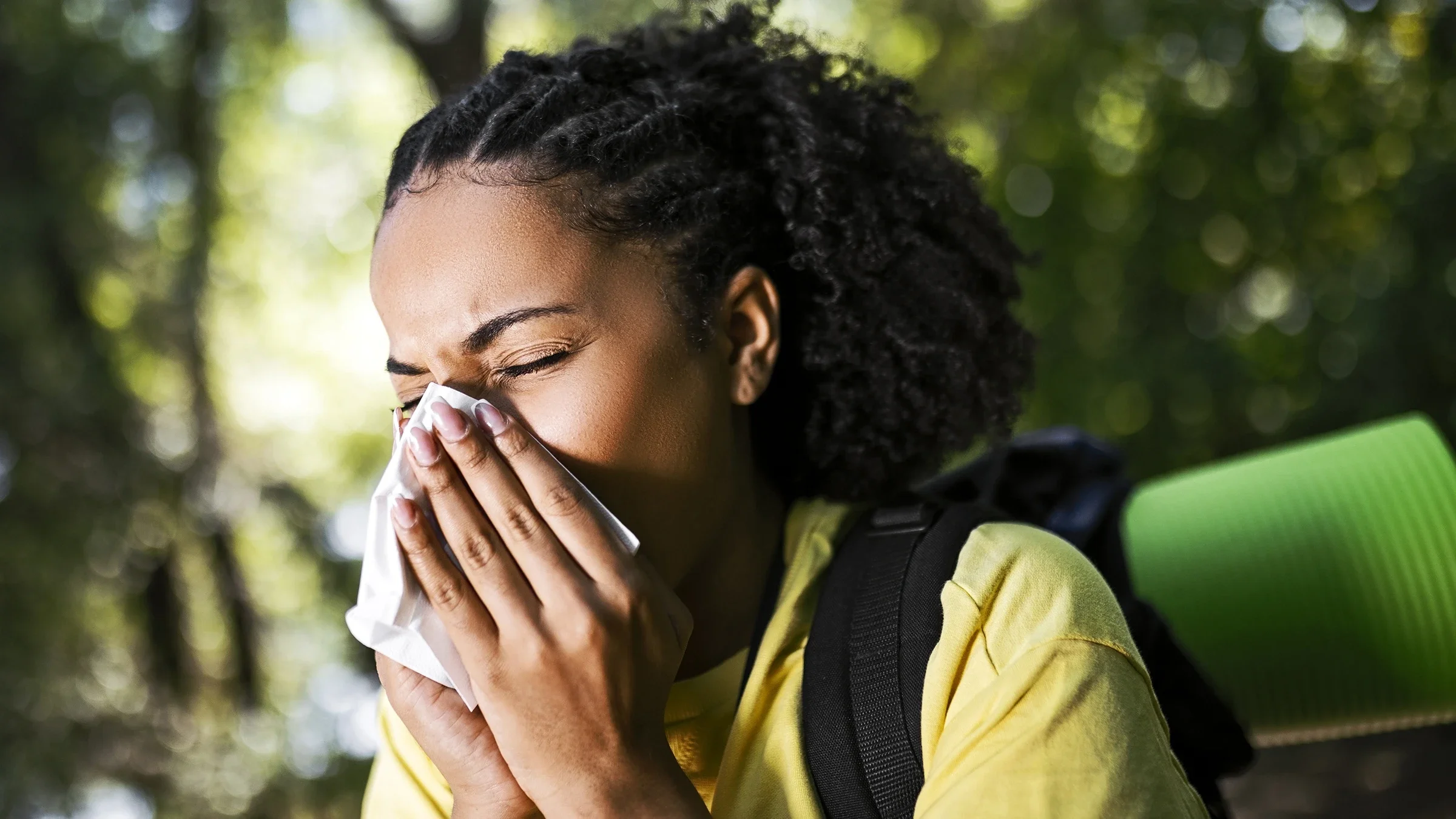 A woman blows her nose while outdoors. Allergies can cause many symptoms, from sneezing to skin rashes.