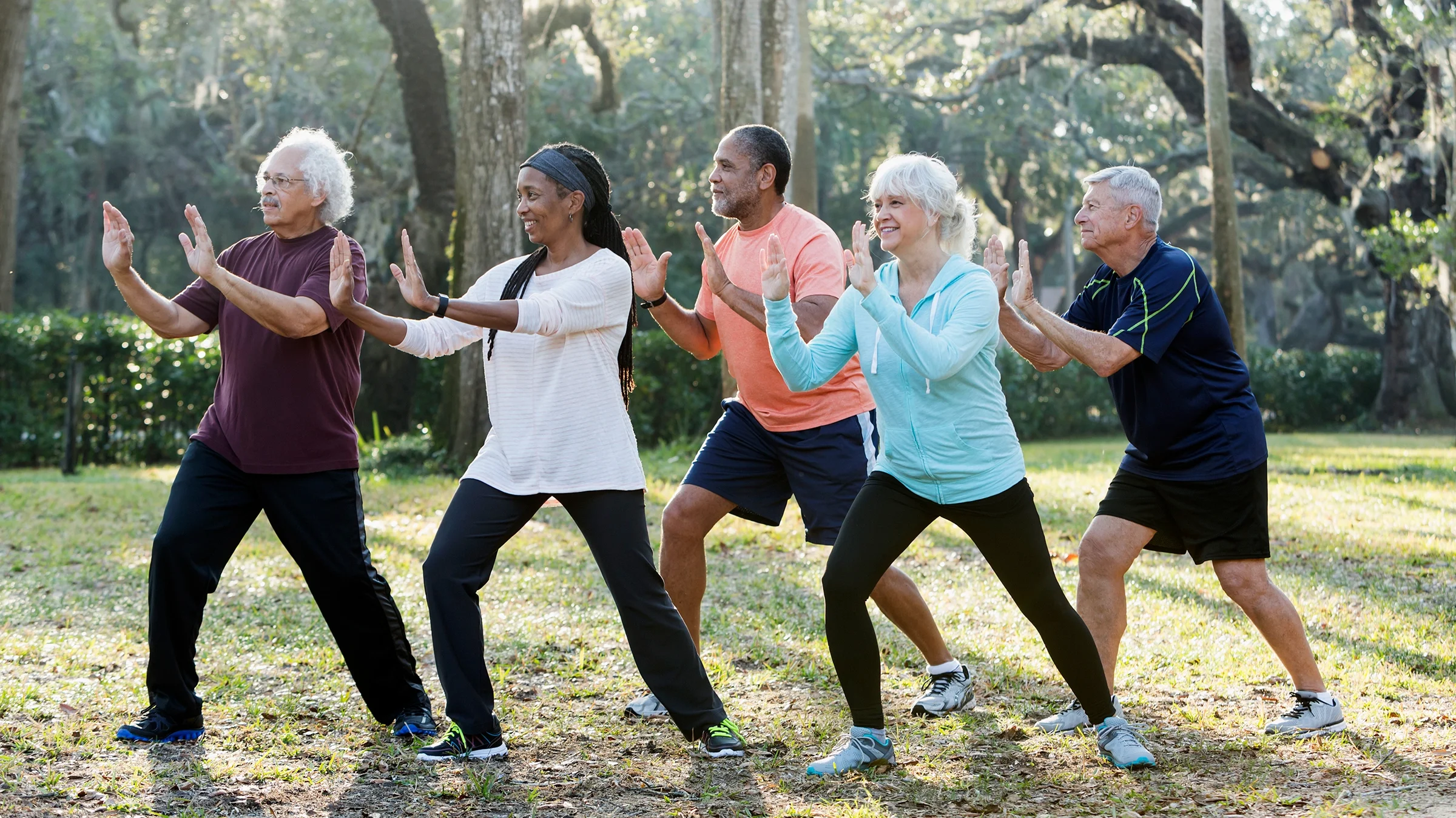 A group of five older people doing tai chi in the park. They are all wearing different types of work out clothes and tennis shoes.