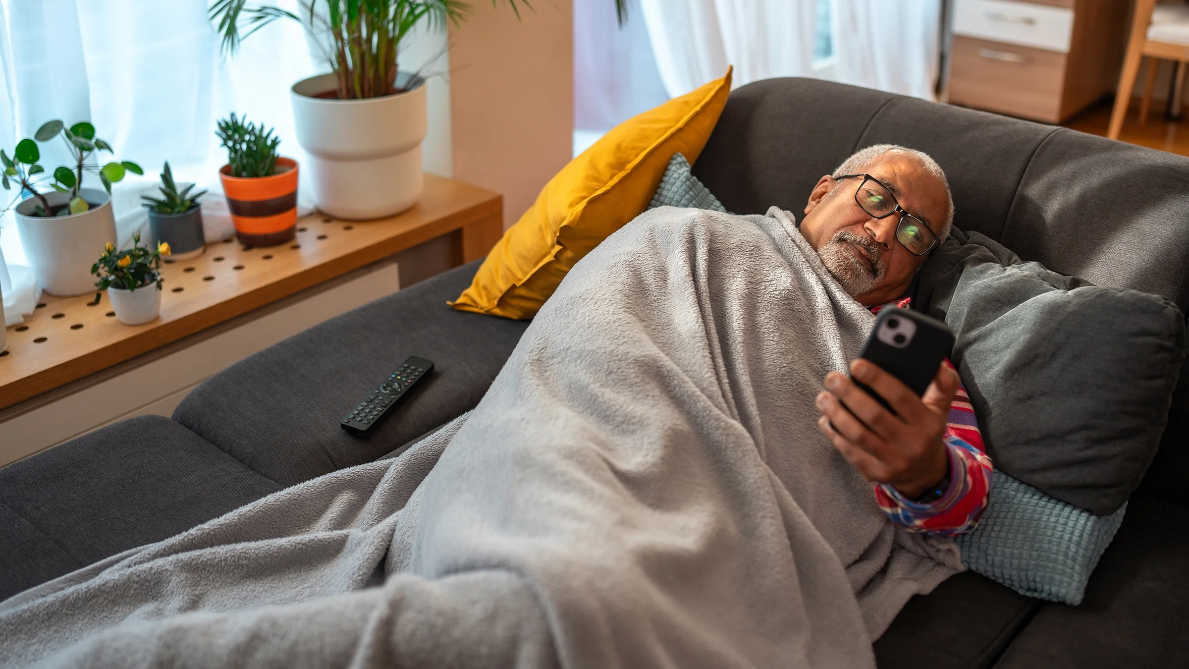 Man lying on a couch under a blanket and reading a smartphone.